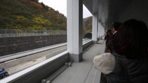Grupo de personas observa desde un andén moderno y cubierto una vía maglev vacía flanqueada por una malla de protección y un talud arbolado con túnel, descripción pensada para accesibilidad y posicionamiento web que incluye la frase Ventajas y desventajas del tren maglev: descubre su impacto y futuro