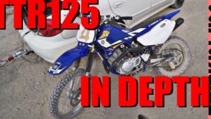 Motocicleta off-road Yamaha azul con plásticos blancos y placa frontal con número, aparcada sobre terreno de grava junto a un coche blanco, con neumáticos de tacos sucios, chasis y motor visibles; Ventajas y desventajas de la TTR 125: ¿es la moto ideal para ti?