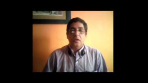 Hombre de mediana edad con gafas y auriculares colgando del cuello, vestido con camisa clara y enmarcado en un plano medio frente a una pared naranja y un cuadro, en una imagen tipo videoconferencia que ilustra Ventajas y desventajas de Twitter en la educación: impacto real.