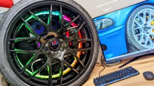Primer plano de una llanta deportiva negra con múltiples radios y luces RGB apoyada frente a un monitor que muestra otra llanta, sobre una mesa con teclado mecánico y ratón, imagen pensada para ilustrar comparativas de hardware y Ventajas y desventajas de un ordenador: descubre qué elegir y por qué