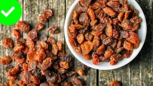 Tazón blanco lleno de uvas pasas de color marrón y ámbar con textura arrugada y brillo natural, varias uvas esparcidas sobre una mesa de madera rústica y un símbolo verde de verificación en la esquina, Ventajas y desventajas de las uvas pasas y sus secretos saludables.