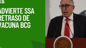 Hombre mayor con traje oscuro, gafas y corbata roja hablando en un atril con el escudo nacional junto a un panel verde con un titular grande sobre el retraso de la vacuna, Ventajas y desventajas de la vacuna BCG: lo que debes saber hoy