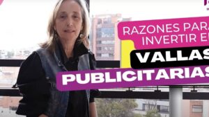 Mujer de mediana edad junto a una ventana con vista a edificios urbanos, señalización gráfica superpuesta en tonos morado, amarillo y negro que promociona invertir en vallas publicitarias destacando el texto visible y el soporte tipo valla, optimizada para SEO con palabras clave sobre publicidad exterior y accesible por alto contraste, y contextualizando 'Ventajas y desventajas de vallas publicitarias y su impacto real'.