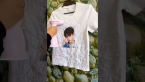 Camiseta blanca colgada con una fotografía impresa en el pecho y abundantemente arrugada siendo alisada con un vaporizador de mano rosa sostenido por una persona, mostrando Plancha de vapor vertical ventajas y desventajas para ti