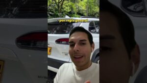 Hombre joven en primer plano grabando un video vertical con auriculares blancos y camiseta clara, sonriendo ligeramente frente a un SUV blanco estacionado entre árboles con una placa amarilla y texto amarillo sobreimpreso 'por regla general', imagen apta para ilustrar Ventajas y desventajas de los zapatos: qué calzado elegir en contenidos sobre estilo urbano y confort.
