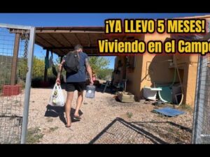Hombre con mochila y bolsas entrando por una verja hacia una vivienda rústica con porche y patio de grava bajo un cielo despejado, imagen evocadora para contenido sobre Ventajas y desventajas de vivir en el campo y la ciudad: qué elegir