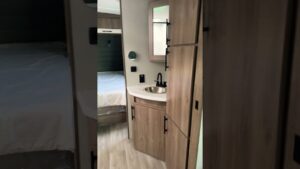 Interior moderno y compacto de un RV con suelo laminado claro, carpintería y armarios de madera, lavabo con espejo y grifo negro junto a una cama con ropa blanca visible al fondo, Ventajas y desventajas de vivir en un RV: descubre si es para ti.