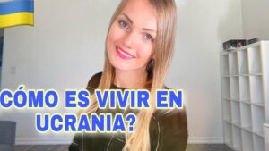 Mujer rubia sonriente en un interior luminoso y minimalista con estantería blanca al fondo, gran texto en azul y amarillo que pregunta cómo es vivir en Ucrania y una bandera ucraniana en la esquina; Ventajas y desventajas de vivir en Ucrania: descubre la verdad real