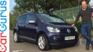 Fotografía de un Volkswagen up compacto de color azul oscuro estacionado frente a una valla metálica, con llantas de aleación y retrovisores plateados y un hombre apoyado en el capó sonriendo, pensada para reseñas y comparativas; Ventajas y desventajas del Volkswagen up: ¿vale la pena comprarlo?