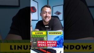 Hombre sonriente sentado frente a la cámara en un escritorio con pósters de componentes en la pared, y en la parte baja dos recuadros comparativos etiquetados LINUX y WINDOWS junto a un cartel rojo que dice DUAL DRIVE DUAL BOOT y un teclado mecánico, imagen ideal para ilustrar Ventajas y desventajas de Windows y Linux: cuál te conviene más