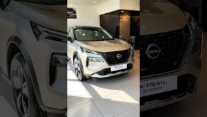Imagen de un Nissan X‑Trail e‑Power plateado exhibido en un concesionario, vista frontal tres cuartos con parrilla V‑Motion y emblema Nissan, faros LED angulares, llantas de aleación y reflejos en el suelo de baldosas que ilustran Nissan X Trail ventajas y desventajas para elegir tu SUV ideal