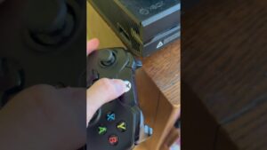 Vista en primer plano de una mano sujetando un mando Xbox negro con el pulgar sobre el botón central y los botones A B X Y visibles junto a una consola Xbox colocada sobre una mesa de madera, imagen ideal para acompañar un análisis titulado Ventajas y desventajas del Xbox One S: ¿vale la pena?