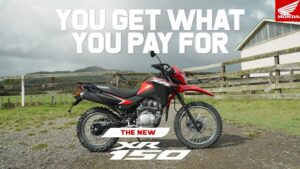 Fotografía de una motocicleta Honda XR 150 en rojo y negro estilo off-road aparcada sobre un camino de grava frente a una cerca de madera y un paisaje rural con cielo nublado, perfecta para descripciones, reseñas y comparativas; Ventajas y desventajas de la XR 150: ¿es la moto ideal para ti?