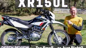 Hombre con equipo de motociclismo amarillo arrodillado en un campo señalando una motocicleta dual-sport blanca Honda XR 150 con énfasis en ruedas, suspensión, motor y ergonomía, imagen pensada para una reseña comparativa Honda XR 150 ventajas y desventajas para elegir la moto ideal.
