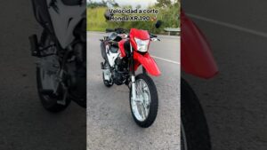 Motocicleta Honda XR 190 en color rojo y blanco, vista en tres cuartos sobre una carretera asfaltada con guardarraíl y vegetación de fondo, con faro delantero rectangular, guardabarros alto y neumáticos mixtos, pensada para búsquedas informativas como Ventajas y desventajas de la XR 190: ¿vale la pena esta moto?