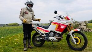 Persona con casco integral y ropa de protección de pie junto a una motocicleta Yamaha de colores blanco, rojo y amarillo con el distintivo "750" aparcada en un prado con flores amarillas y cielo nublado, Ventajas y desventajas de la Yamaha XTZ 125 para comprar