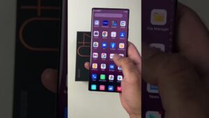 Vista frontal de un ZTE Axon 50 Lite sostenido con la mano sobre un fondo claro que muestra la pantalla de inicio con múltiples iconos de aplicaciones, bordes curvados y la caja del teléfono en segundo plano, Ventajas y desventajas del celular ZTE Axon 50 Lite: ¿vale la pena?