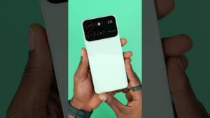 Manos sujetando un smartphone blanco visto por detrás con un prominente módulo de cámara rectangular de tres lentes y logo ZTE sobre fondo verde, con muñeca con reloj y dedo con anillo, Zte Blade A3 2020 ventajas y desventajas para decidir tu compra