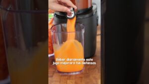 Vaso transparente que se llena con zumo de naranja de color brillante vertido desde un extractor doméstico sobre una encimera, mostrando la textura y pulpa del jugo y un mensaje sobre mejorar las defensas, Ventajas y desventajas del zumo de naranja: ¿es saludable?